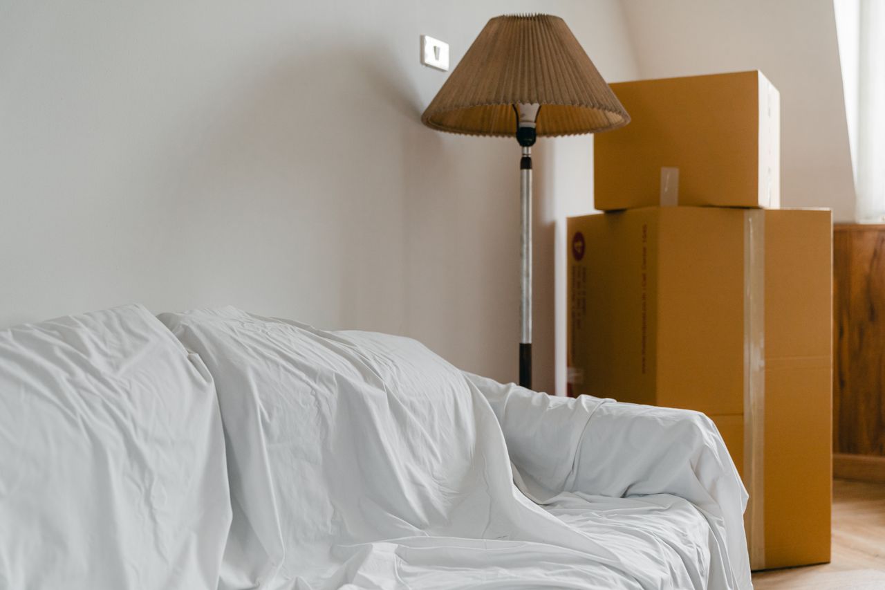 Canapé recouvert d’un drap, lampe sur pied et cartons emballés dans une pièce lumineuse et spacieuse d’une maison contemporaine neuve